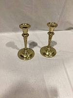 Two brass candlestick holders side by side on a white cloth background, showing front and base edges.