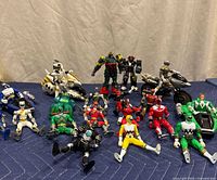 Photo showing multiple Power Rangers action figures and toy vehicles arranged on a blue cloth. Figures include red, green, white, yellow, black, and other Ranger suits. Several motorcycles and a small vehicle are behind them.