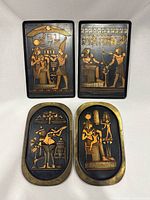 Four ancient Egyptian art metal wall plaques displayed against white background, two rectangular and two oval shaped, showing front detailed design in gold-tone and black enamel or paint.