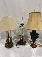 Three mixed material lamps, two candle-style brass lamps with off-white pleated lampshades and one brass marbled brown lamp with beige lampshade. All on a white backdrop showing cords and tarnish.