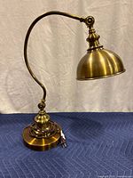 Full view of the brass gooseneck desk lamp showing the curved arm and round base with coiled cord and power plug.