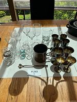 Overall view of 24-piece lot of vintage stemware and glassware on table includes etched crystal wine glasses, brass goblets, silver plated cup, and swizzle stick.