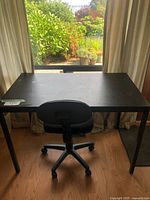 Photo of black wooden rectangular desk with four legs and black office chair placed in front, showing scratches and wear on desk surface.