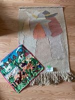 Vintage woven wall hanging with three abstract figures wearing hats, beige background with fringe bottom, alongside a colorful embroidery piece depicting sheep and rural scenery.