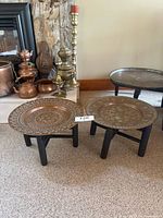 Two small vintage Middle Eastern copper tray tables with black wooden bases. The copper tops have intricate engraving in floral and geometric patterns in silver detail on copper. One base looks unstable and needs repair.