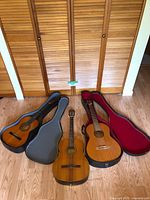 Three guitars displayed on floor: grey soft gig bag case on left, central guitar upright without case, red-lined hard case on right