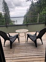 Two black plastic Muskoka chairs and small light-colored round table on wooden deck overlooking water view on cloudy day.