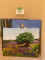 Three acrylic paintings stacked against a wall including a large tree in a wildflower field, with two other nature scenes partially visible behind it