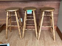 Front view of three solid wood bar stools with round seats and four legs, placed side by side on wood floor against wood panel wall.