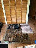 Three woven textiles laid out on a wood floor in front of louvered closet doors: two large tapestries or rugs featuring abstract, geometric, and figurative designs, and one smaller striped woven piece with fringe.