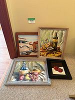 Four paintings on floor leaning against a wall including still life and scenic art.