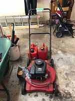 Photo of a red Briggs & Stratton lawn mower with black engine and three red jerry cans next to it in a garage.