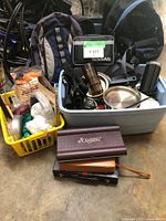 Two backpacks, kitchen utensils, thermos, yellow basket with toiletries, Scrabble game and other travel items on the ground.