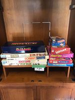 Stack of classic board games on wooden shelf including Pictionary, Clue, Sorry, Monopoly, Pay Day, Boggle, Yahtzee, Pass the Pigs, and Rummikub.