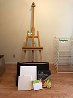 Photo showing full wooden easel with adjustable clamp and canvas shelf, black art portfolio bag in front, canvas pad and blank canvas boards displayed on bag.