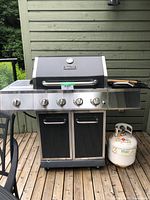 Front view of Dyna-Glo propane grill with five burner control knobs, attached propane tank on side, and grill accessories on side shelf.