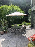 Full view of outdoor patio set showing glass table, four chairs with cushions, and beige umbrella in a garden background.