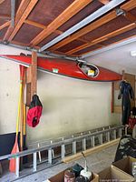 Red one-person kayak suspended horizontally from ceiling mounts in garage with spray skirt attached to cockpit opening.