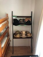 Leaning black metal frame shelf with two wooden shelves holding various hats near bunk bed.