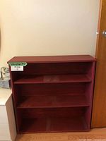 Maroon painted wooden bookshelf with three shelves, positioned against a wall near a door.
