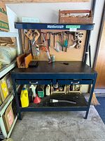 Front view of the Mastercraft workbench showing black worktop, pegboard with tools, and lower shelves with containers.