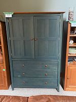 Front view of a solid wooden armoire painted dark green with a natural wood top, two doors, and three drawers with wooden knobs.
