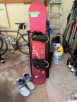 Photo of entire snowboard standing upright with bindings and snowboard boots on the floor in front, showing overall condition and setup.