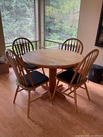 Full view of round oak pedestal table with four wooden chairs and black chair pads positioned in a room with wood floor and large windows.