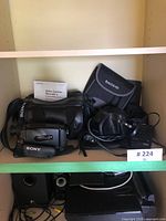Photo showing Sony Handycam Video8 camcorder with Sony shoulder strap, original manual, carrying bag, Tasco binoculars in soft case, Panasonic camera in leather case, and battery charger with cords.
