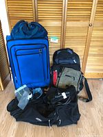 Wide view of five travel bags including a large blue suitcase, backpacks, a shoulder bag, and hydration pack against wooden closet doors.