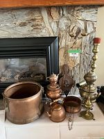 Wide view of displayed items on table against a stone fireplace wall: large copper boiler, brass samovar, copper teapot, small copper pot, wooden paddle with ship carving, and brass decorative piece.