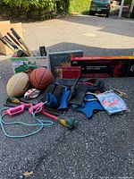 Assorted sports balls, badminton rackets in box, jump rope, and neoprene gloves laid on asphalt.