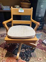 Full view of vintage oak desk chair with beige cushion on a patterned rug in office setting.