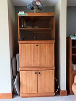 Front view of vintage wood storage unit showing glass display shelf on top, drop-down bar shelf middle section, and closed lower cabinet doors.