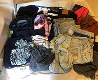 Wide view of the clothing lot showing folded jeans, tops, sweaters, and jackets mostly in dark and neutral colors with some colorful tops; items appear lightly used.
