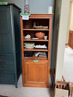 Front and slight angle view of the tall wood storage unit placed next to a dark green cabinet, showing three open shelves and one closed cupboard door at the bottom, no items included.