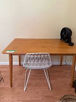 Rectangular wood table with mid-century modern style tapered legs, light-medium wood finish shown from a front view with a white wire chair in front of it and a small black fan on the table.