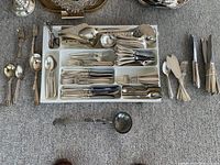 White tray filled with assorted silver cutlery including forks, knives, and spoons, plus additional serving utensils arranged around it.