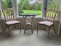 Two rattan chairs with floral cushions and a round rattan table with glass top set up near a window.
