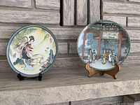 Two collector plates displayed on stands against a stone background showing detailed traditional Chinese artwork with female figures.