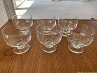 Photo showing six clear glass dessert cups arranged in two rows of three, on a wooden surface with outdoor background visible.