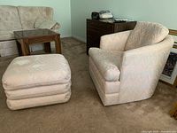Pink and grey tub chair and matching cushioned stool on brown carpet, with other furniture in background.