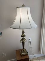Tall brass table lamp with classic design and white fabric bell-shaped lampshade, visible electrical cord plugged into wall outlet.