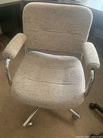 Close-up of the tan fleck fabric office chair showing padded seat, backrest, and armrests with chrome metal base and wheels.