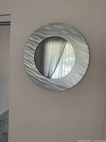 Front view showing the round mirror with a wide brushed aluminium frame with wavy texture reflecting window curtains.