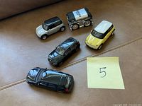 Photo of five assorted toy cars on a couch including two Mini Cooper-style models, a black SUV, a dark blue sedan, and a toy shaped like a cassette tape.