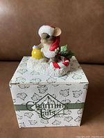 Ceramic mouse figurine on top of its original Charming Tales box, dressed in a red Santa hat, holding a yellow ornament ball, with a candy cane and holly decoration on the base.