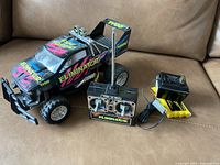 Photo of a black electric remote control pickup truck with 'Eliminator' decals, a black wired remote controller, and a yellow battery charger with battery pack, showing the entire lot contents on a brown couch.
