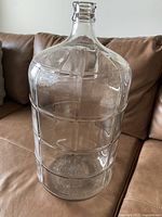 Clear glass carboy displayed on brown leather couch, showing full body and neck details with no visible damage.