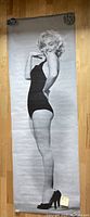 Full view of the life size Marilyn Monroe poster showing her in a black swimsuit standing sideways with black heels on wooden floor, measuring approximately 77x26 inches.